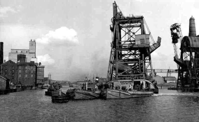 On Whit Monday, 2 June 1952, the floating No 4 Compartment Boat Hoist is being towed back to its berth in Goole's South Dock, following dredging operations. At least two Aire and Calder Navigation tugs, with their jebus' in front, are towing the structure. Also in view is No 5 Compartment Boat Hoist. Since this photo was taken the hoist was 'listed' as Grade 2* and is of national importance.    Harold Crabtree Collection